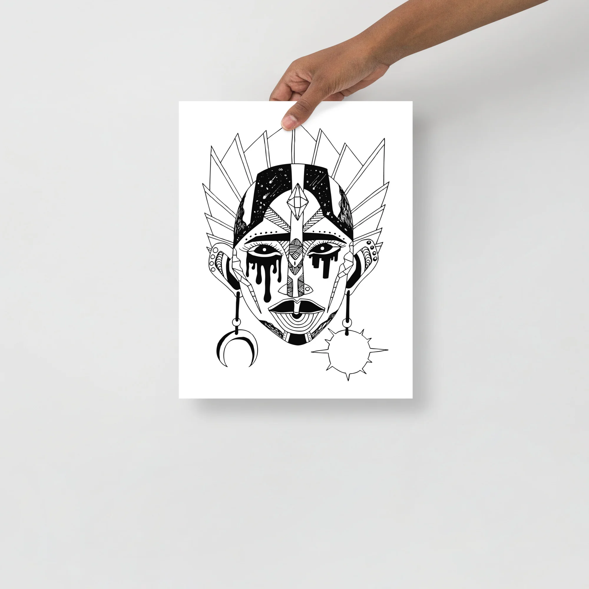 Black and white African mask art print featuring an ornate tribal face with geometric patterns, crescent and sun symbols, held up by hand