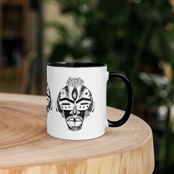 White ceramic African mask coffee mug with black handle and intricate geometric tribal face design featuring decorative patterns and crown-like crown detail.