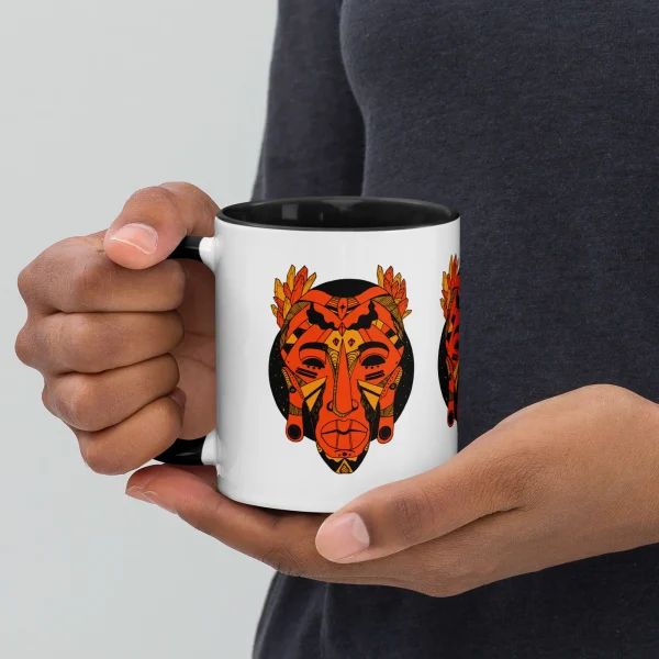 Person holding white ceramic african mask coffee mug with orange and black tribal art design