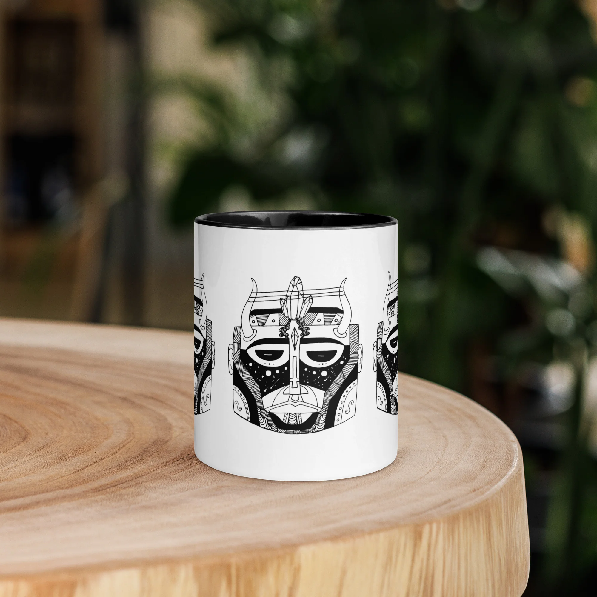 White ceramic African mask mug with black intricate tribal design and black interior rim, displayed on wooden table with green foliage background.