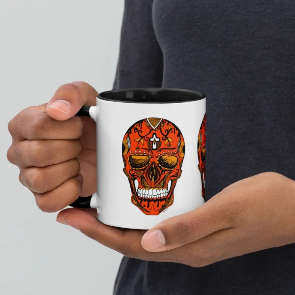Hand holding a day of the dead coffee mug with bold orange sugar skull design and gold accents on white ceramic.