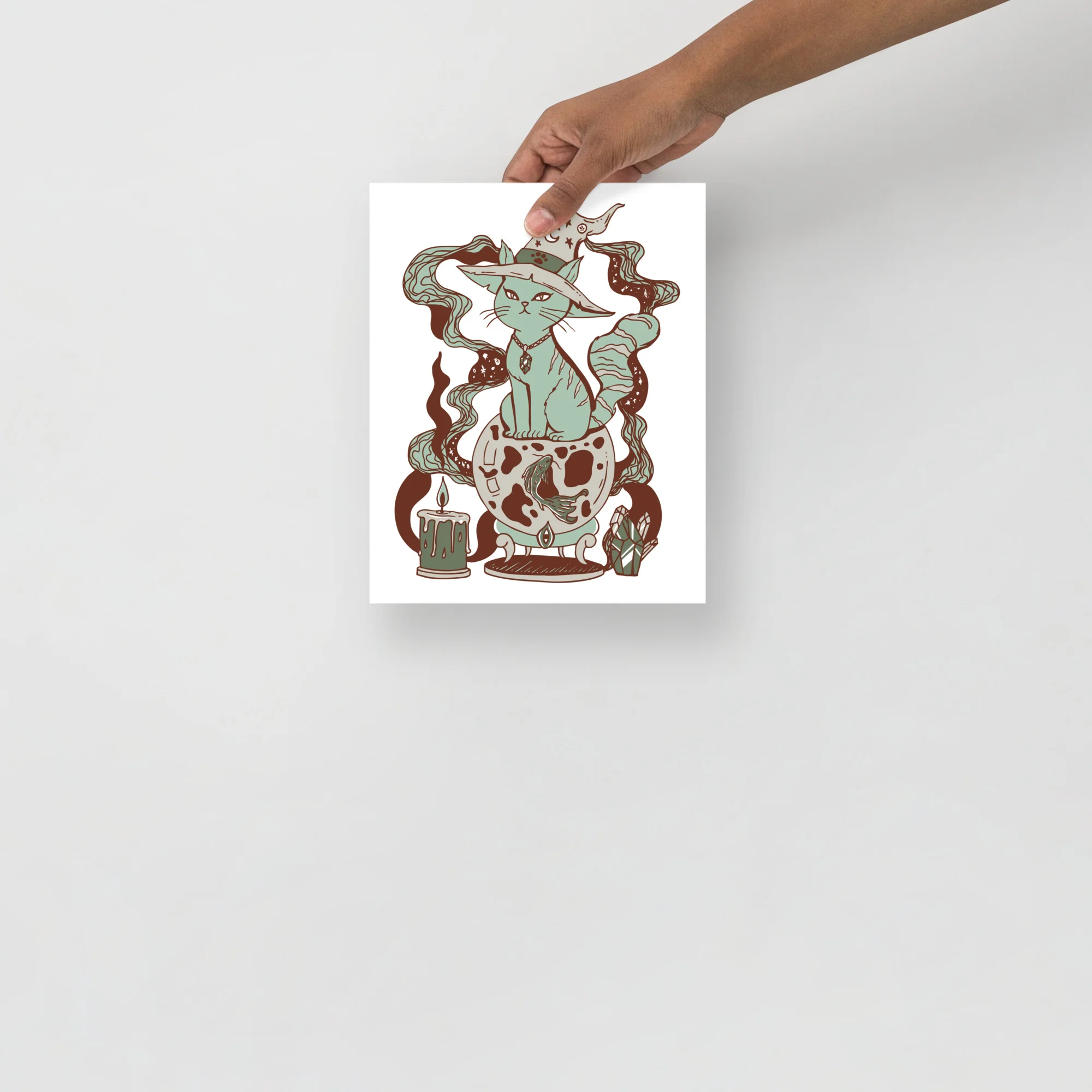 Sage green cat art print held in hand showing intricate line-drawn design with mystical elements and earthy brown accents on white matte paper.
