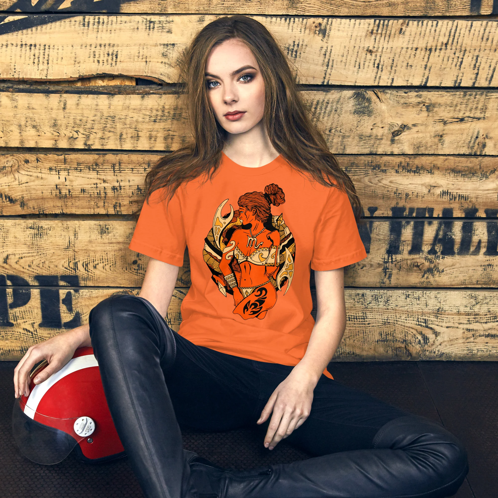 Woman wearing an orange scorpio t-shirt with black warrior artwork and gold accents, seated against wooden backdrop